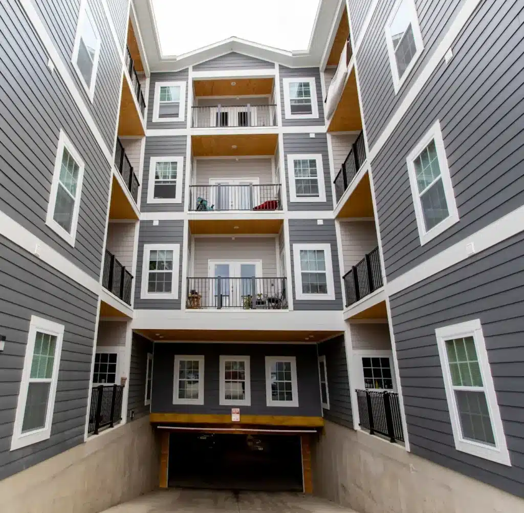 Four-story apartment building with gray siding, white trim, balconies, and a ground-level entrance to an underground parking garage.