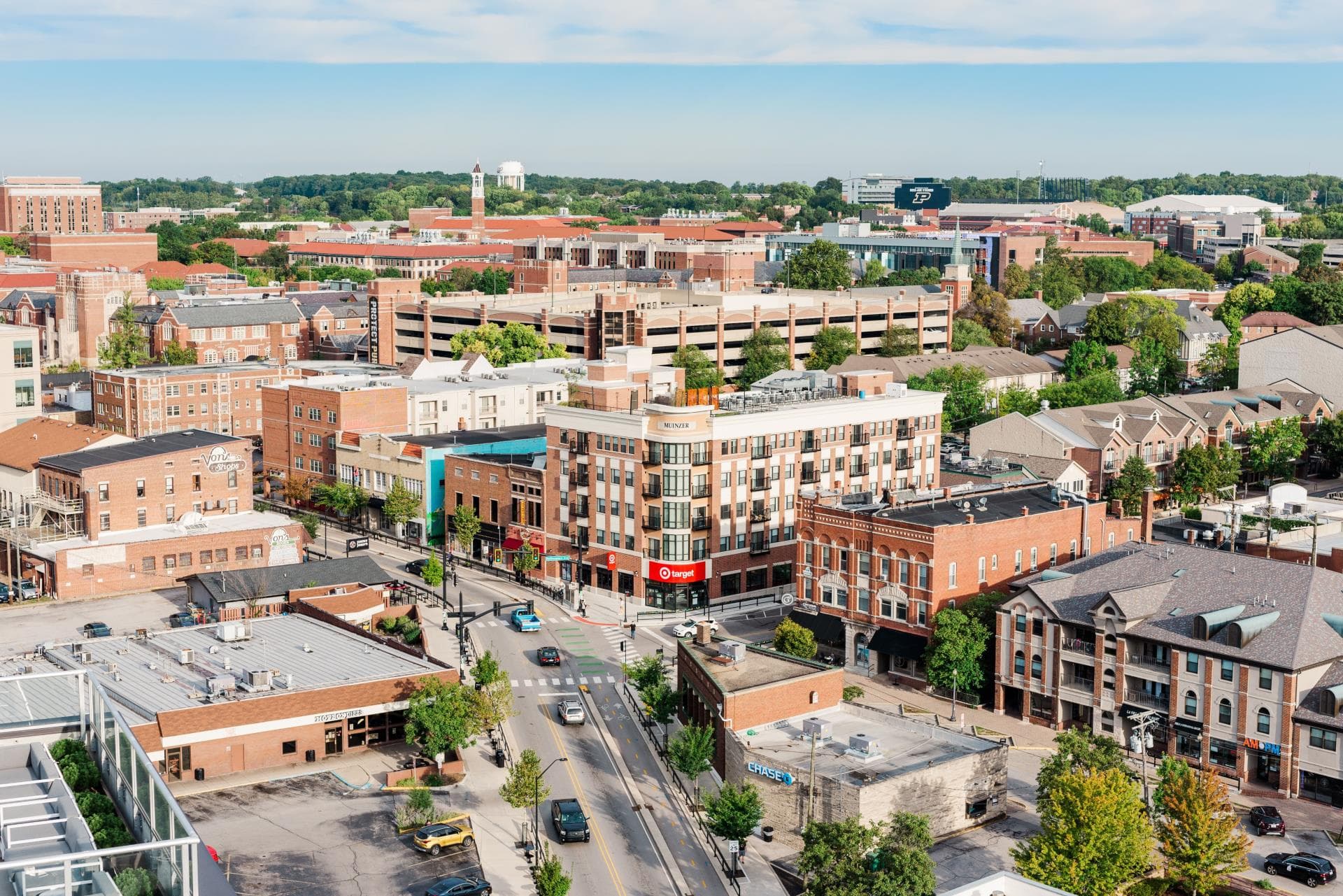 Aerial view of an urban area with brick buildings, a parking garage, roads, trees, and a university campus in the background under a partly cloudy sky.