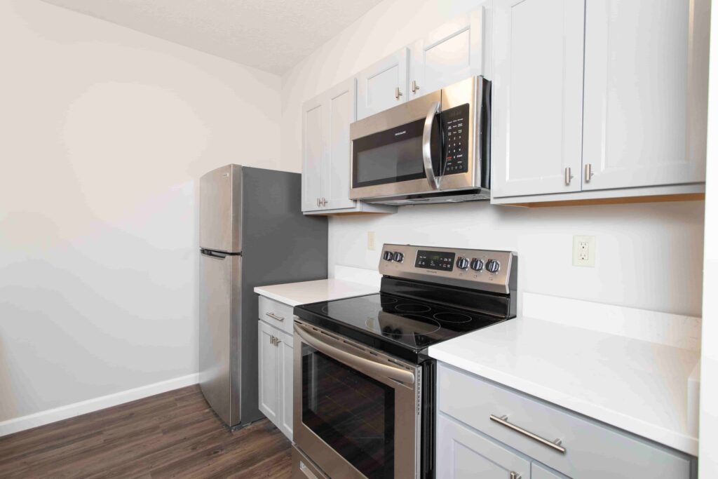 Modern kitchen with stainless steel appliances, including a refrigerator, oven, and microwave, white countertops, light gray cabinets, and wood-look flooring.