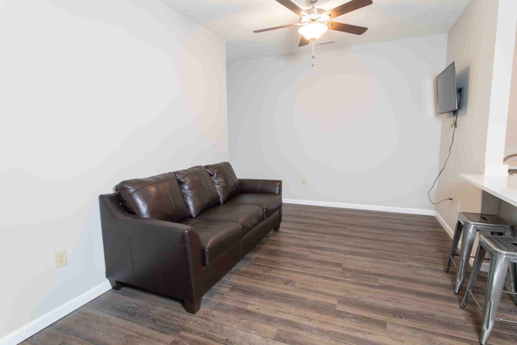 A small living area with a dark brown leather sofa, ceiling fan, wall-mounted TV, and wood-look flooring. Three metal barstools are beside a white counter on the right.