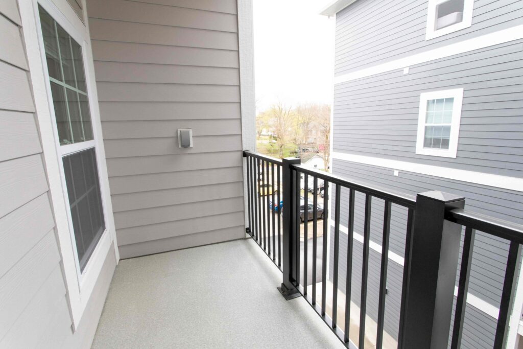 A small balcony with a light gray floor, black metal railing, and beige siding, overlooking a street and neighboring gray building.
