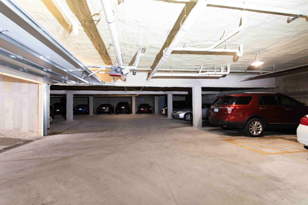 Underground parking garage with several parked cars, concrete floor, exposed ceiling pipes, and fluorescent lighting.