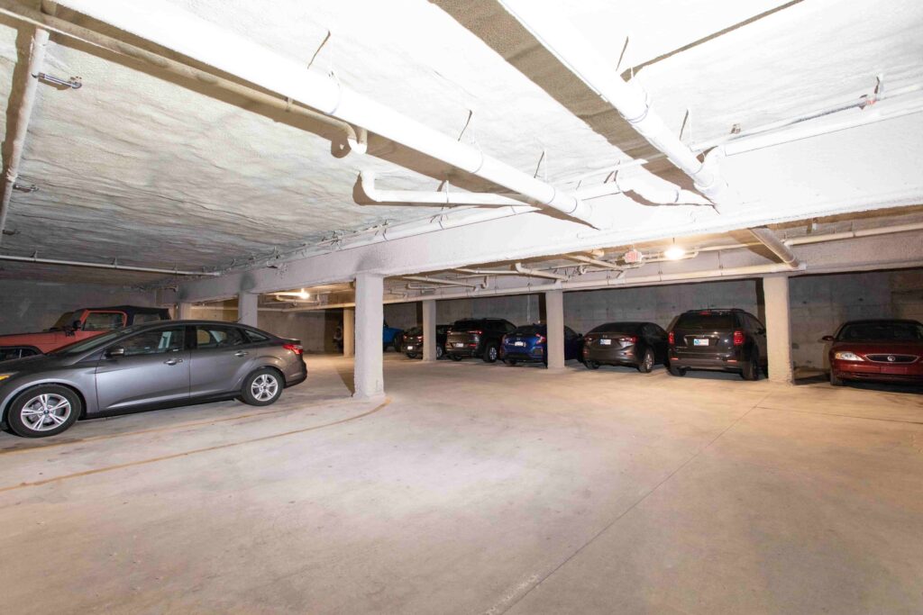 Indoor parking garage with concrete columns and several parked cars along the right side, illuminated by overhead fluorescent lighting.