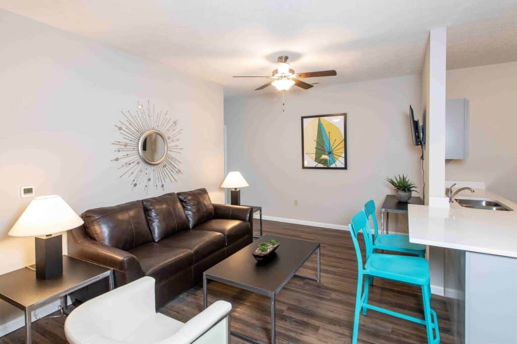 Modern living room with a brown leather sofa, two tables, wall art, decorative mirror, two turquoise bar stools, lamps, and a ceiling fan. Kitchen counter visible on the right.