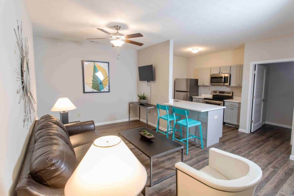 Modern apartment living room and kitchen with gray cabinets, stainless steel appliances, a brown leather sofa, white armchair, and turquoise bar stools at the island.
