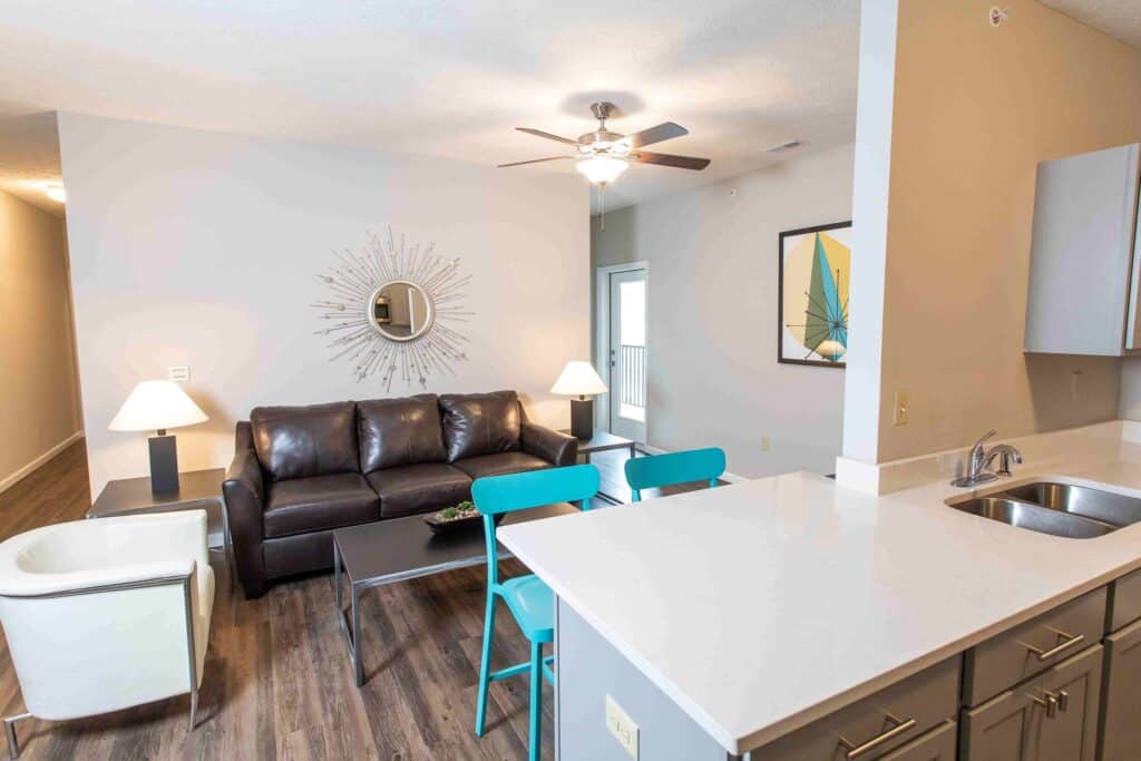 Modern living room with a dark leather sofa, white armchair, coffee table, two turquoise chairs, ceiling fan, wall mirror, and kitchen area with white countertop and sink.