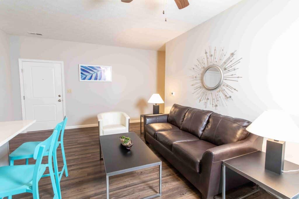 A living room with a brown leather sofa, white armchair, black coffee table, turquoise chairs, and modern decor, including a sunburst mirror and table lamps.