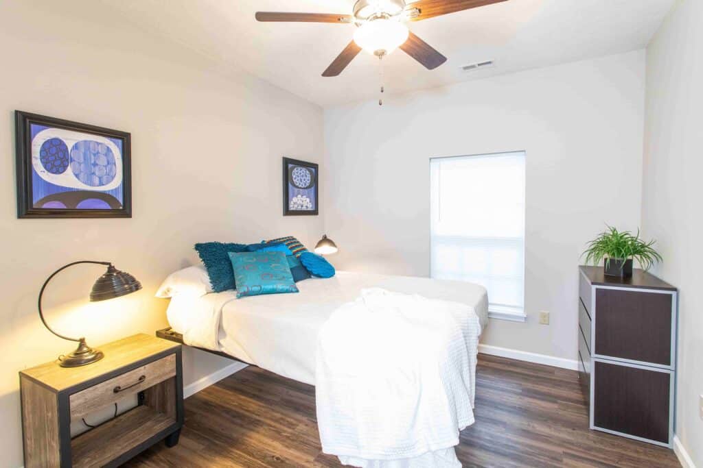 A small, bright bedroom with a bed, blue accent pillows, a nightstand with a lamp, a dresser, wall art, and a ceiling fan.