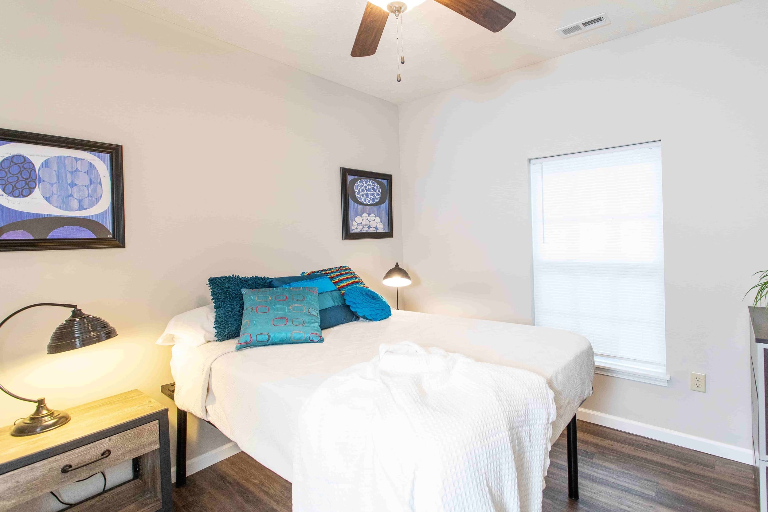 A neatly made bed with blue accent pillows, a nightstand with a lamp, framed artwork on the wall, and a window with closed blinds in a bright bedroom.