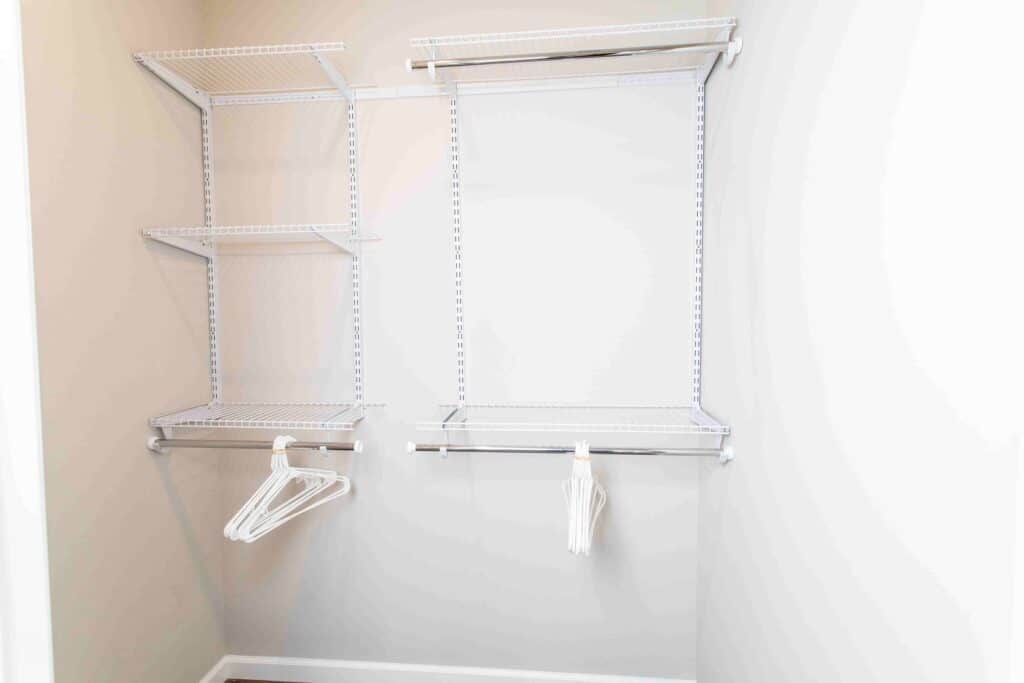 Empty closet with white wire shelves and hanging rods, holding a few white plastic hangers, against plain light-colored walls.