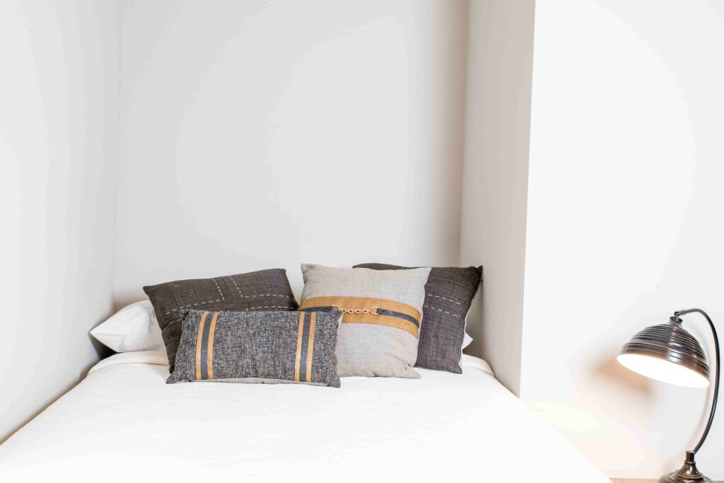 A bed with four decorative pillows is placed in a corner next to a floor lamp with a black shade.