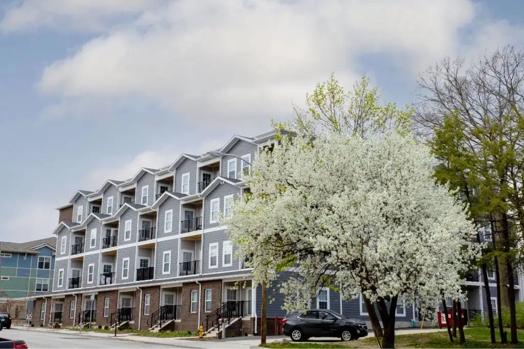 A four-story apartment building with gray siding stands beside a blooming white tree and parked cars under a partly cloudy sky.