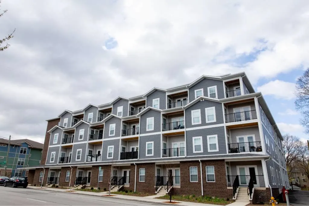 A four-story modern apartment building with brick on the lower level, gray siding above, and multiple balconies, photographed under a cloudy sky.