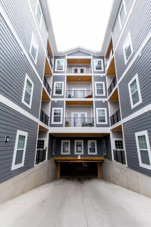 Four-story gray apartment building with white trim, featuring balconies and a driveway leading to a covered parking area at the ground level.
