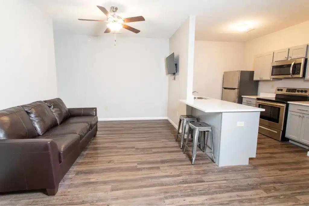 A modern apartment kitchen and living area with a brown leather sofa, wood flooring, a ceiling fan, stainless steel appliances, and a breakfast bar with metal stools.