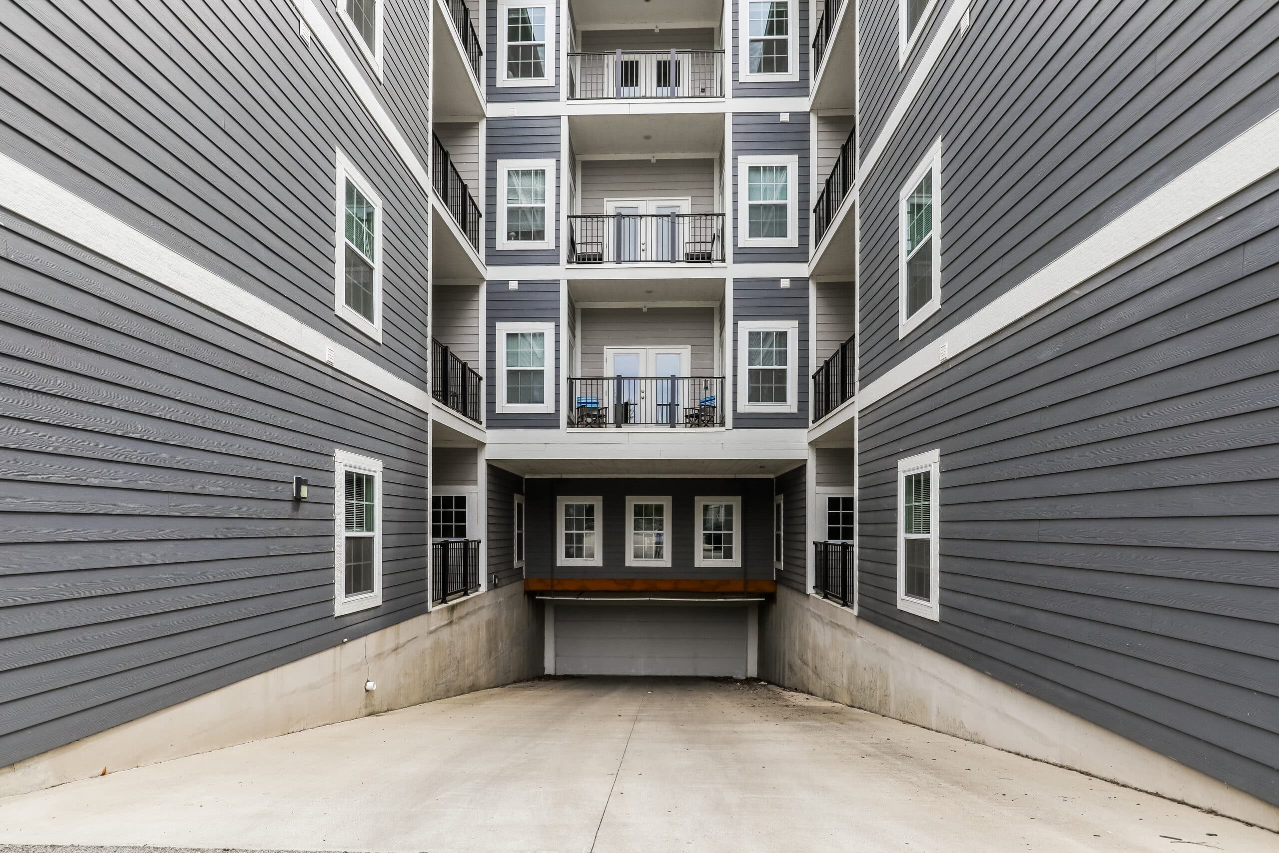 Gray apartment building with white trim, multiple balconies, and a driveway leading to a covered parking area at ground level.